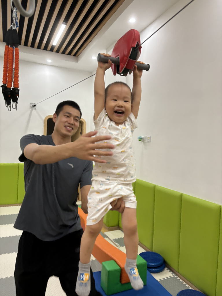 A joyful child hanging from gym equipment with the support of an adult in a padded playroom.