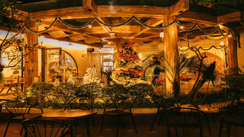 Cozy outdoor cafe at night, illuminated with warm string lights and decorated Christmas trees.