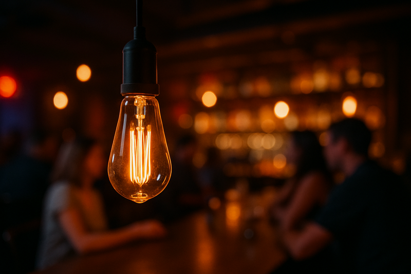 A close-up of an LED Edison bulb hanging over a bar counter, with blurred background of patrons enjoying themselves.